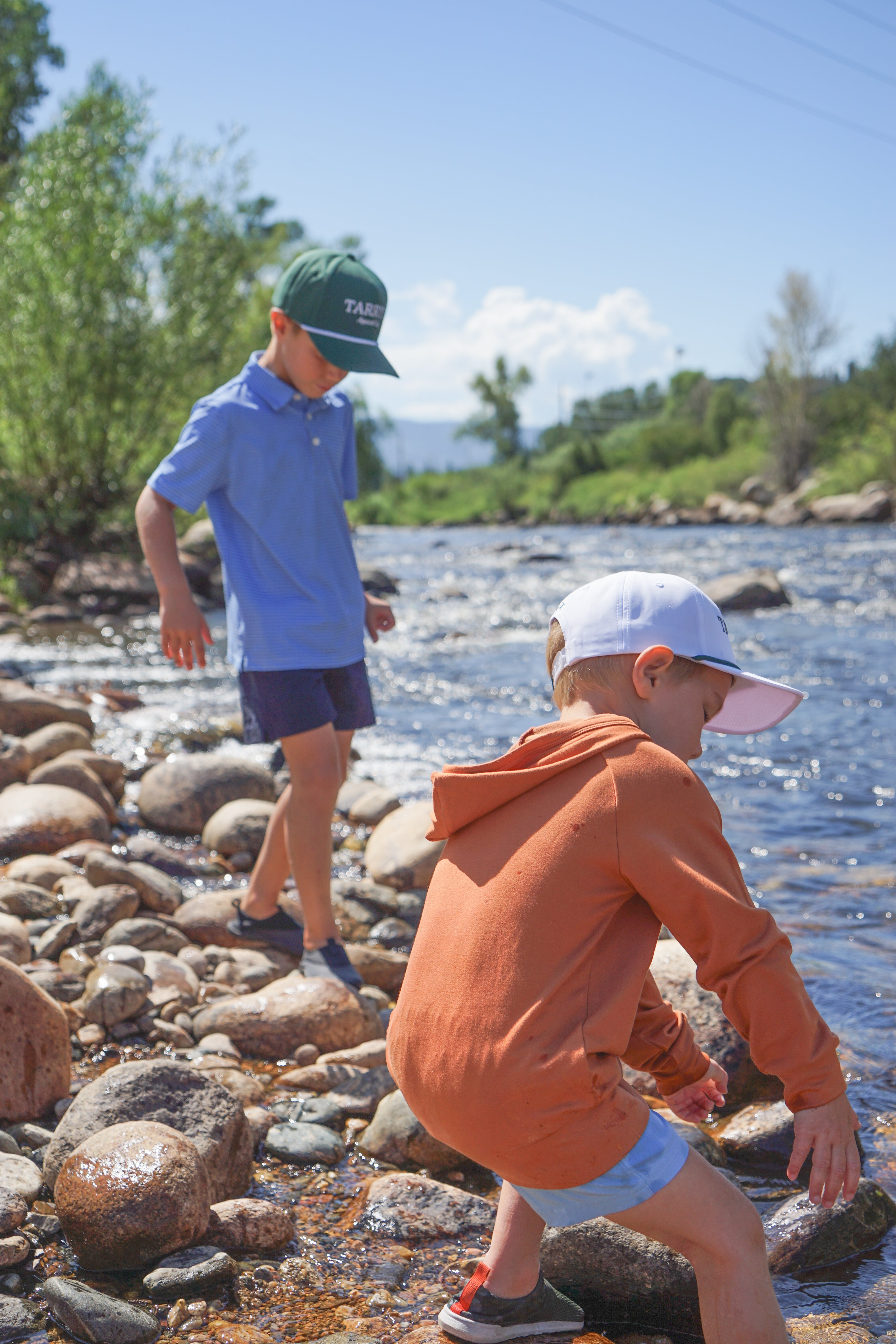 Two boys river with rocks and water in the background wearing blue and navy polo and burnt orange hoodie
