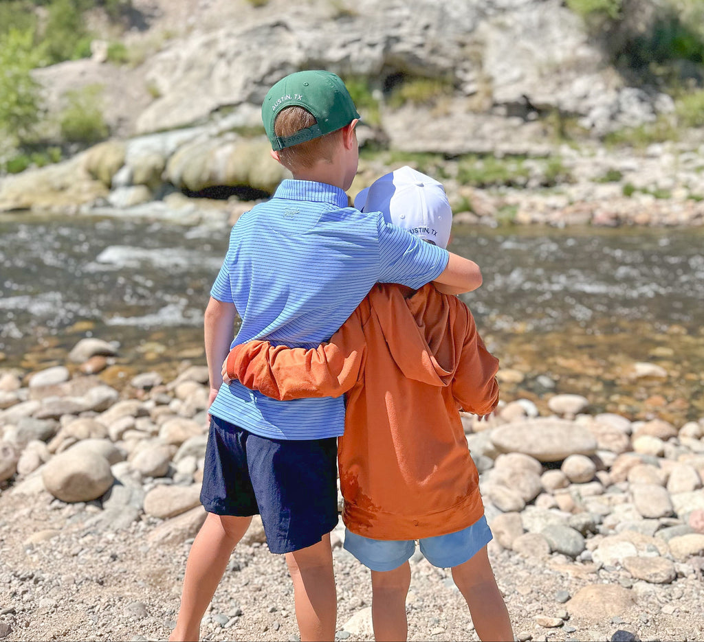 Two boys hugging by a river with rocky landscape in blue and navy polo and burnt orange hoodie