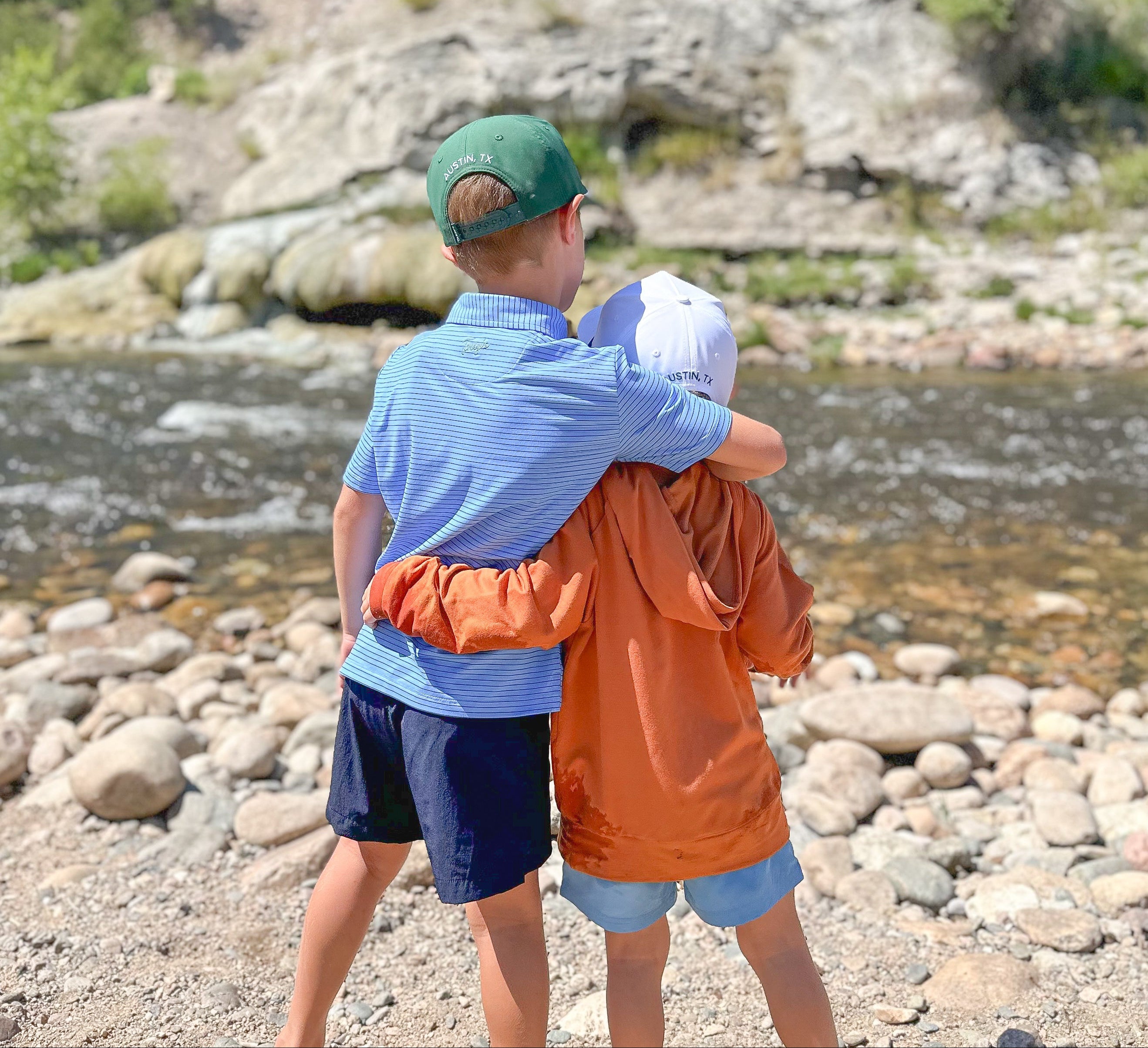 Two boys hugging by a river with rocky landscape in blue and navy polo and burnt orange hoodie