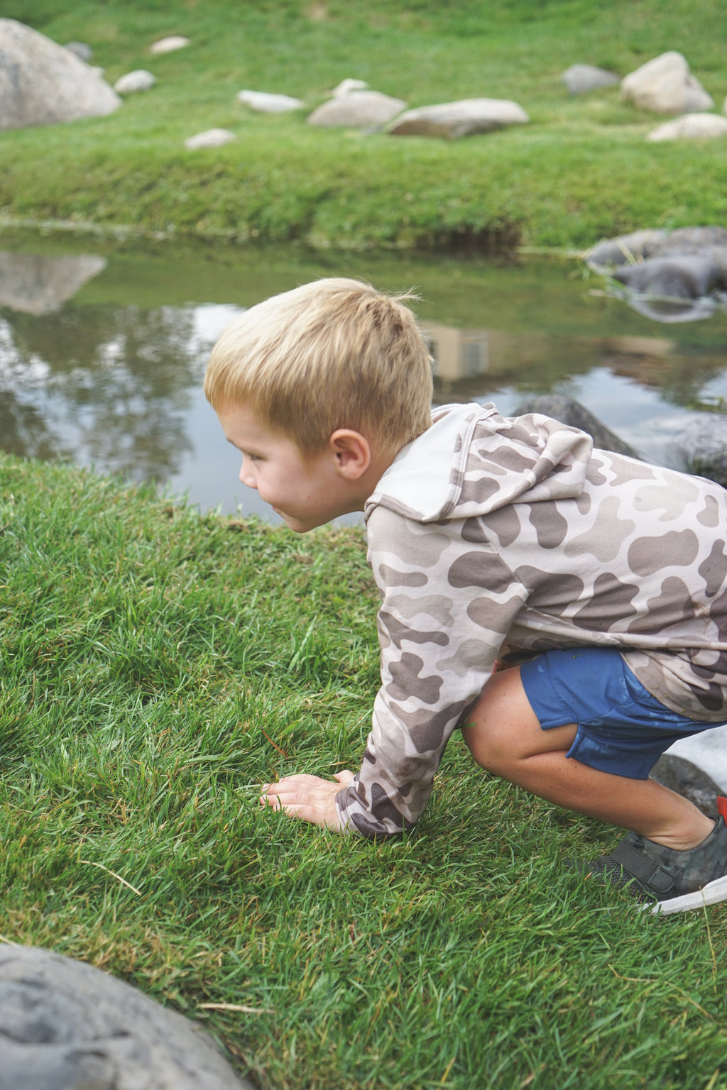 Boy in camouflage hoodie and blue shorts crouching by a pond