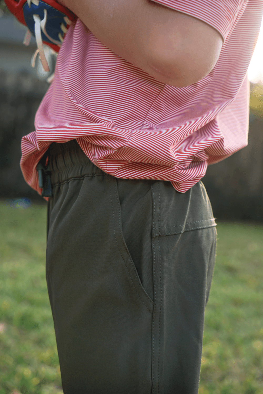 Boy wearing forest green pants and a red and white striped polo standing in a grassy field