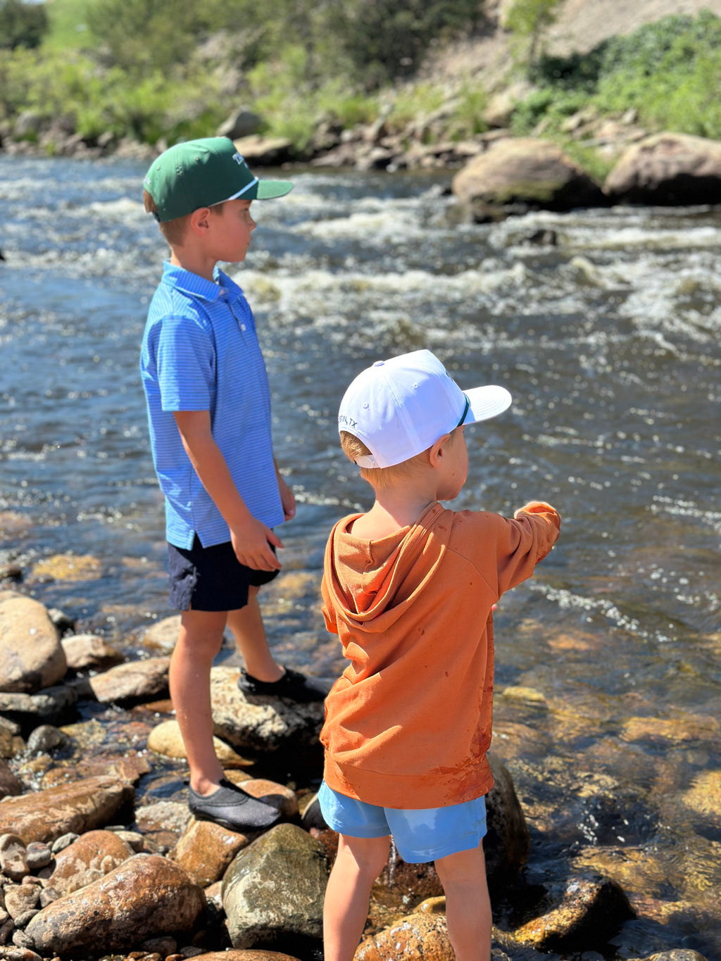 Two children standing by a river, one wearing a blue shirt and green cap, the other in an orange hoodie and white cap.